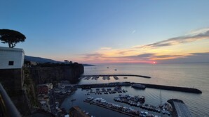 Ein Küstenhafen mit vertäuten Booten, ein Sonnenuntergang im Hintergrund und ein Gebäude am Fels.