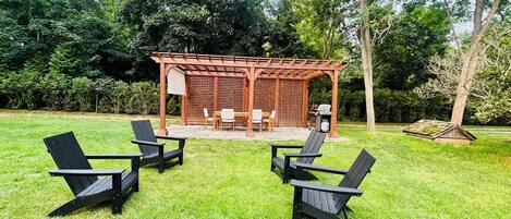 A gazebo with a table and chairs, surrounded by a grassy lawn with Adirondack chairs.