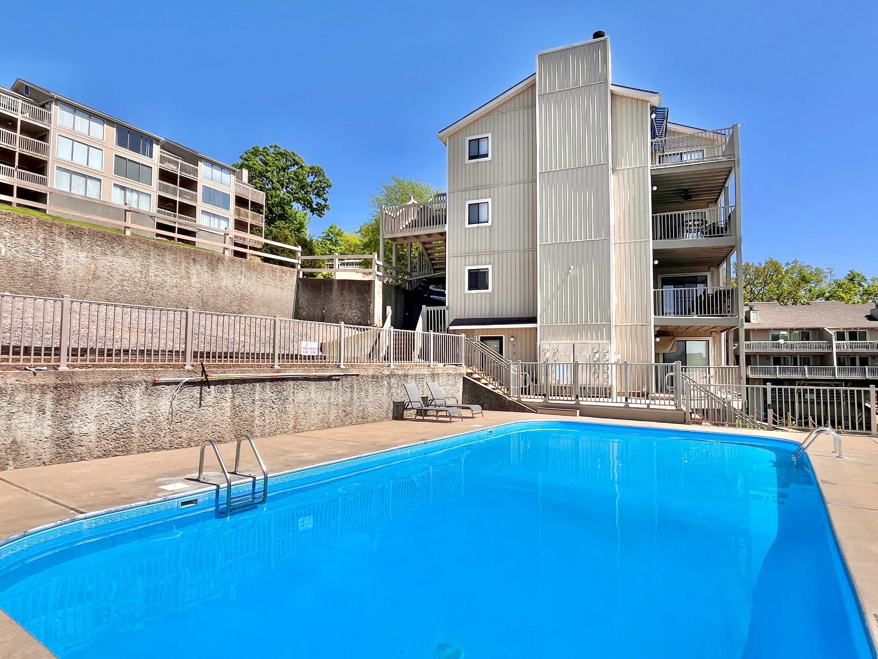 En swimmingpool med en metallstege, belägen intill en hög byggnad med flera våningar, balkonger och en mur av sten som fungerar som stödmur.