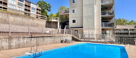 En swimmingpool med en metallstege, belägen intill en hög byggnad med flera våningar, balkonger och en mur av sten som fungerar som stödmur.