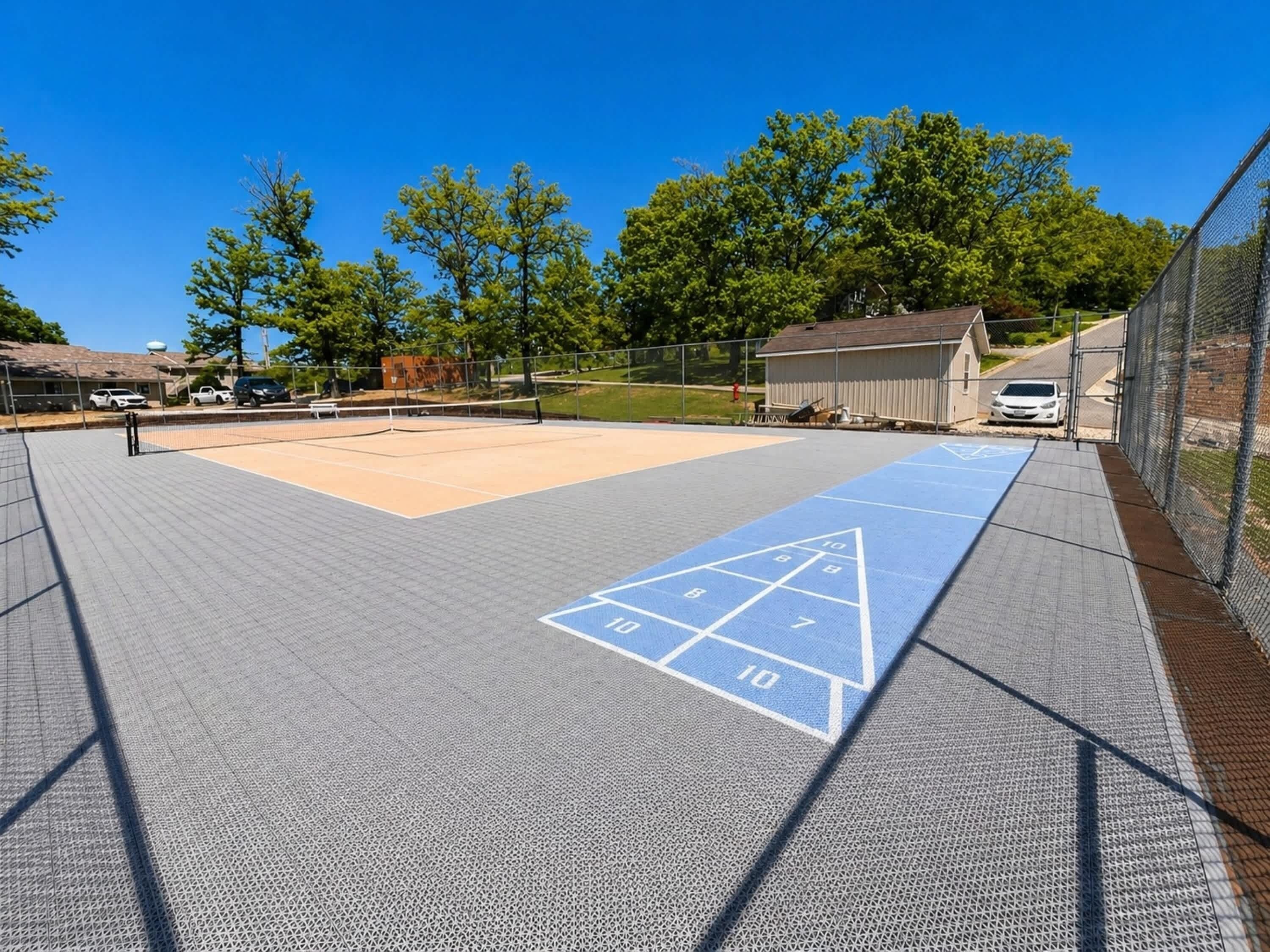 A tennis court with a shuffleboard area, surrounded by a fence, trees, and a building in the background.