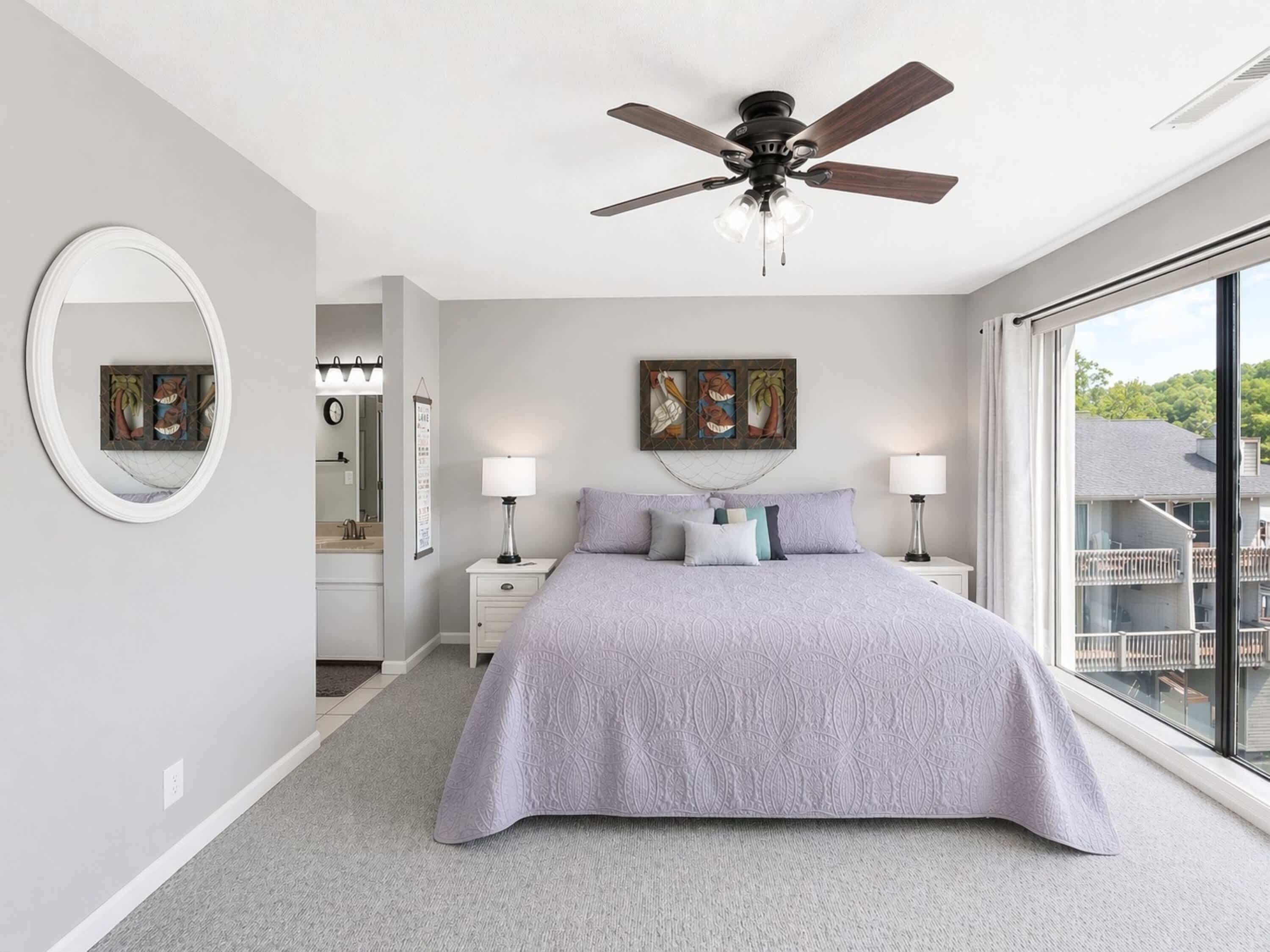 A bedroom with a bed, two bedside tables with lamps, a ceiling fan, a large window, and artwork on the wall.