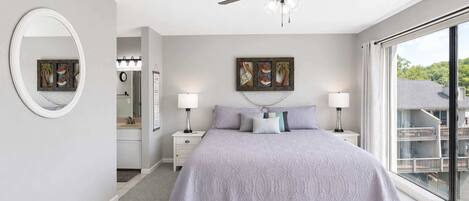 A bedroom with a bed, two bedside tables with lamps, a ceiling fan, a large window, and artwork on the wall.
