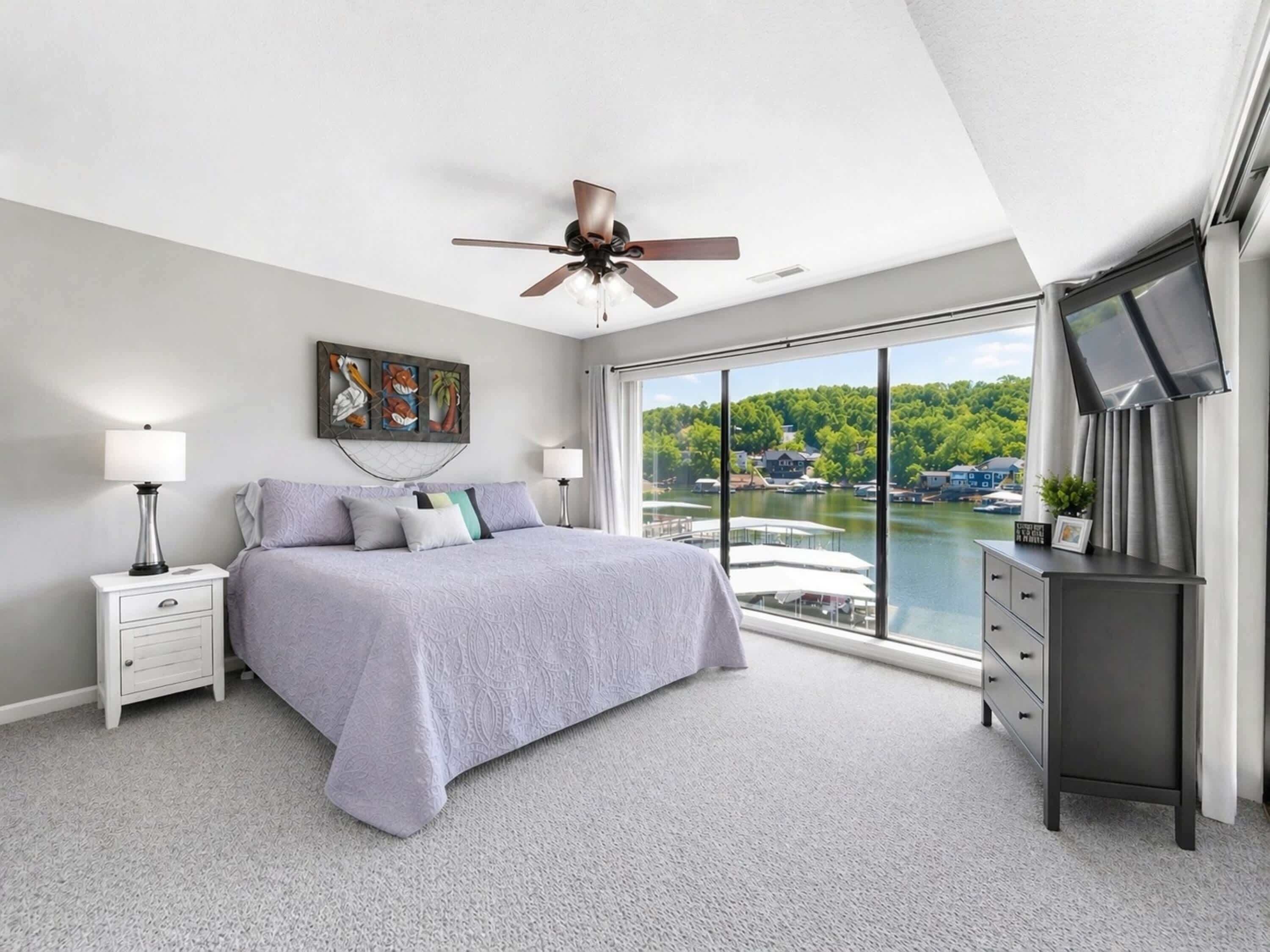 Une chambre avec un grand lit, un ventilateur de plafond, une table de chevet, une commode et une vue sur un lac.