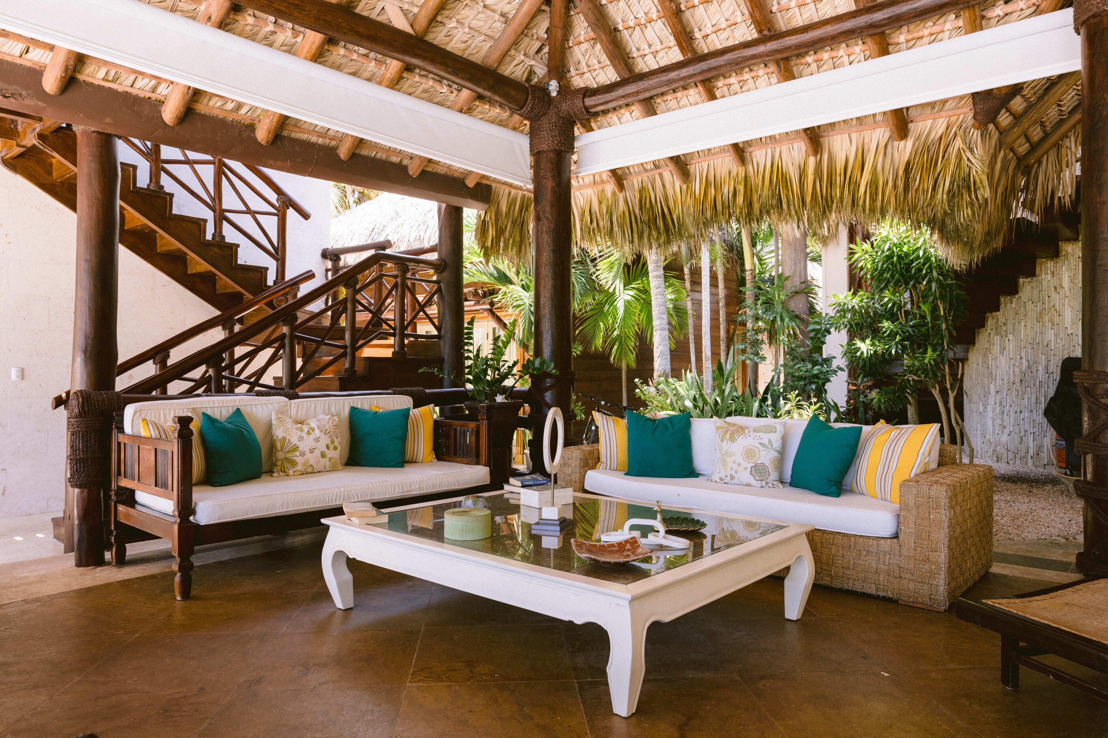 A spacious indoor area with a wooden staircase, two sofas with cushions, a glass coffee table, and a thatched roof structure.