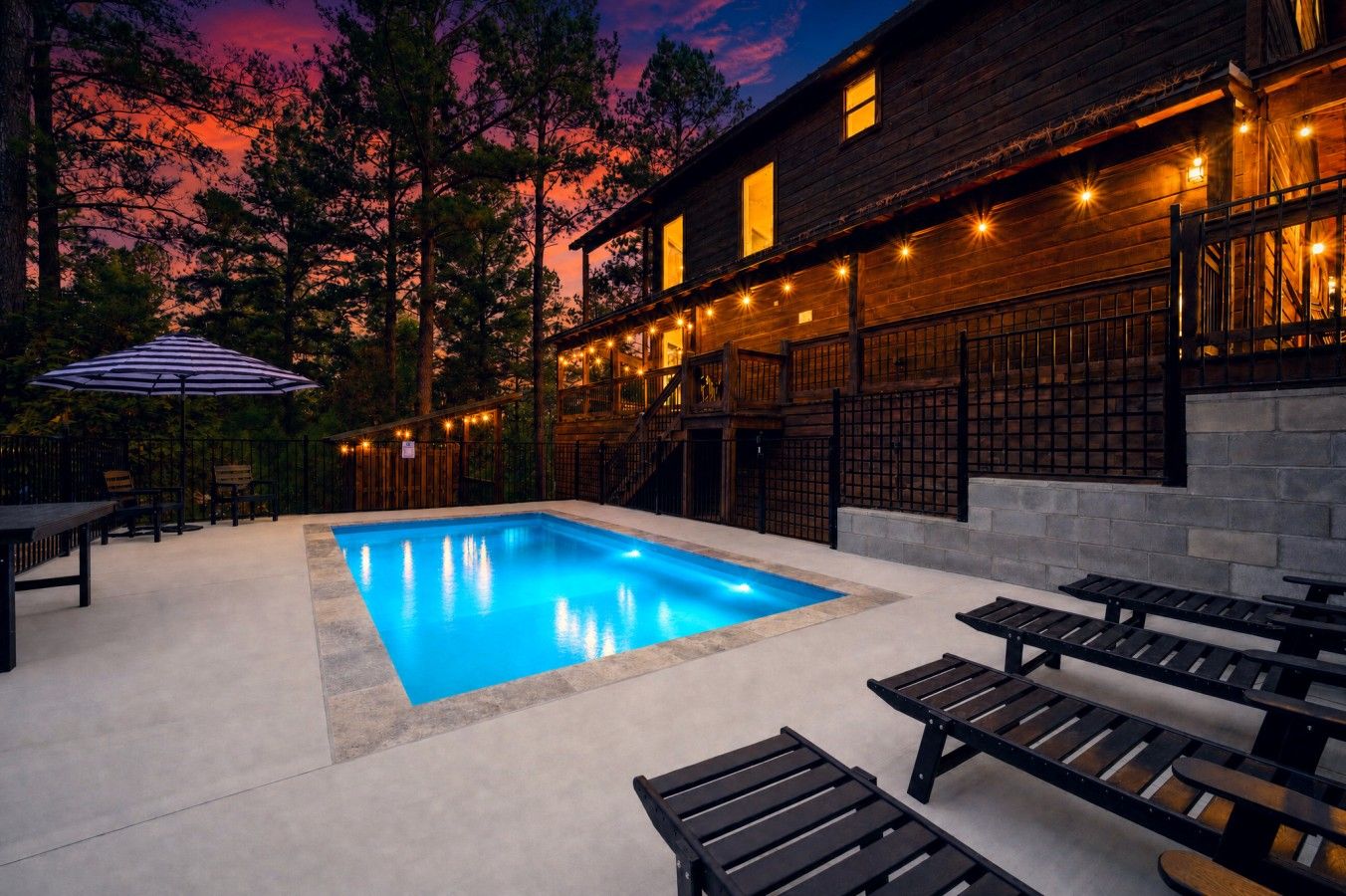 A swimming pool with a wooden deck, outdoor seating, and a building with string lights.