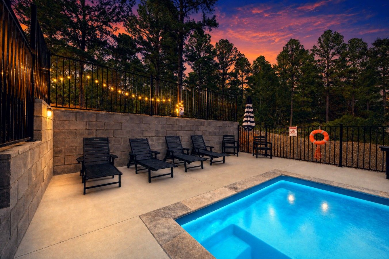 Lounge poolside under string lights as the sky fades from orange to deep purple.
