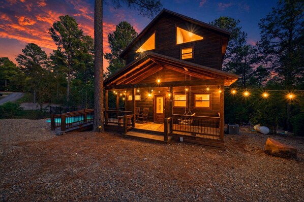 A two-story wooden cabin with a porch, a pool, and string lights. - Blue Water Pines · New! Heated Pool · Ping Pong, Hot Tub and Fire Pit (Broken Bow)