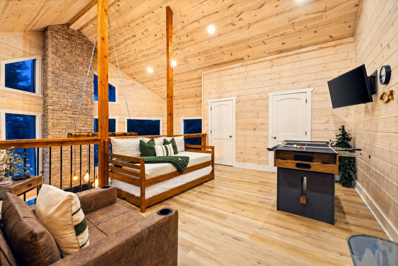 A cozy room with a wooden ceiling, a bed, a desk with a foosball table, and a TV on the wall.