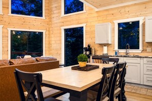 Ein modernes Interieur mit einem Essbereich aus Holz, einer Küche mit weißen Schränken und großen Fenstern, die einen Blick ins Freie ermöglichen.