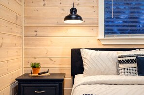 Ein Schlafzimmer mit einer Holzwand, einem schwarzen Nachttisch mit Pflanze, einer weißen Bettdecke und einem Fenster mit Jalousien.