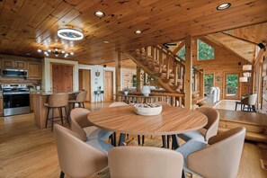 A spacious wooden interior with a round dining table, bar stools, a staircase, and a kitchen area. - Serene Mtn Cabin Retreat,  Lake Views , Hot Tub (New Tazewell)