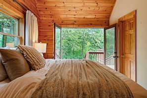 Un dormitorio de cabaña de madera con una cama, almohadas, una lámpara y vistas al bosque a través de puertas abiertas.
