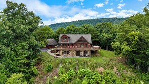 Une grande cabane en rondins à deux étages, dotée d’un balcon, entourée d’une forêt dense et de collines.
