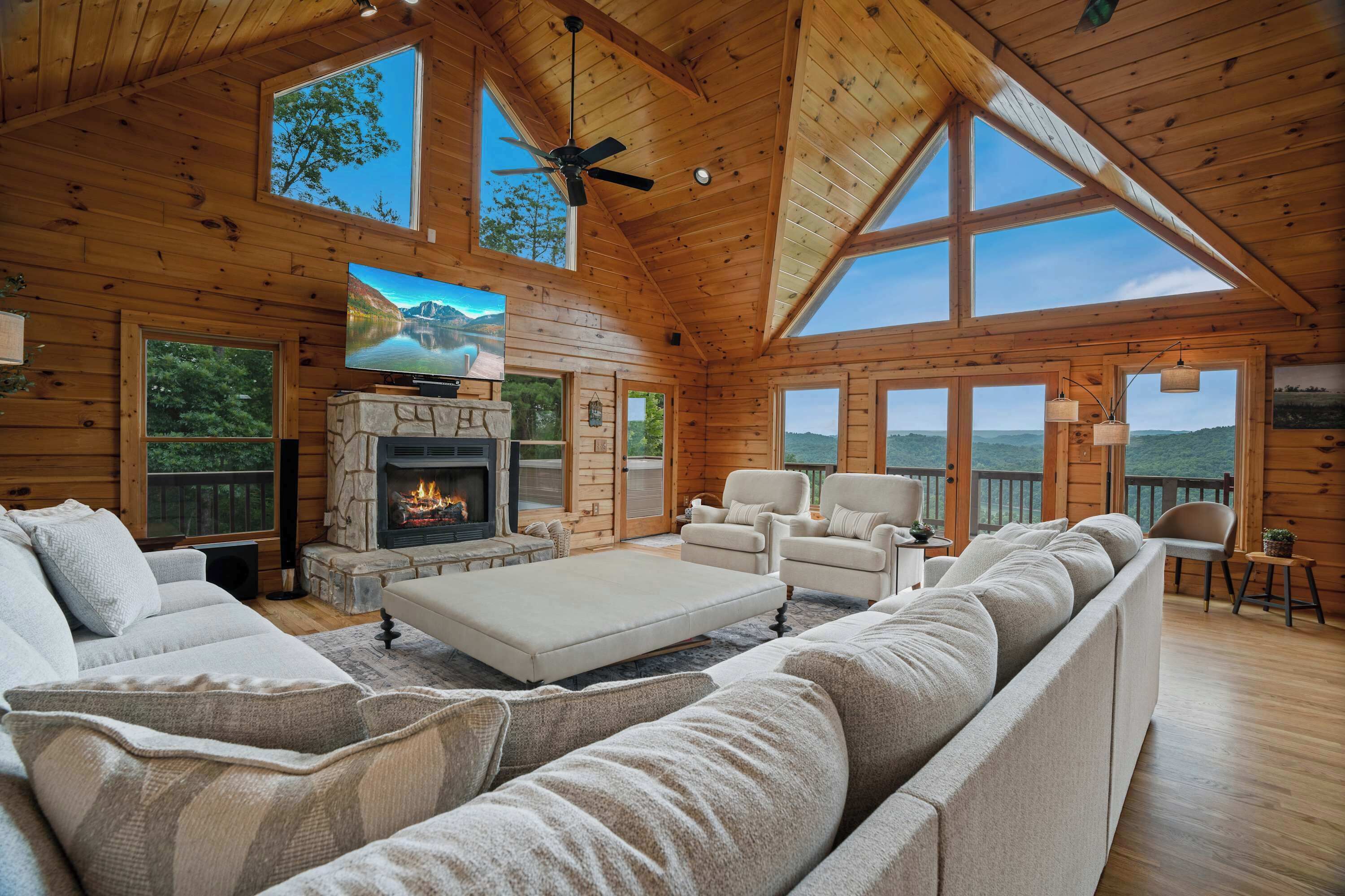 A log cabin interior with a fireplace, a large TV, a sofa, and a ceiling fan.