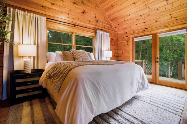 Un dormitorio de cabaña de madera con una cama, dos lámparas de noche, una ventana y vistas a árboles.