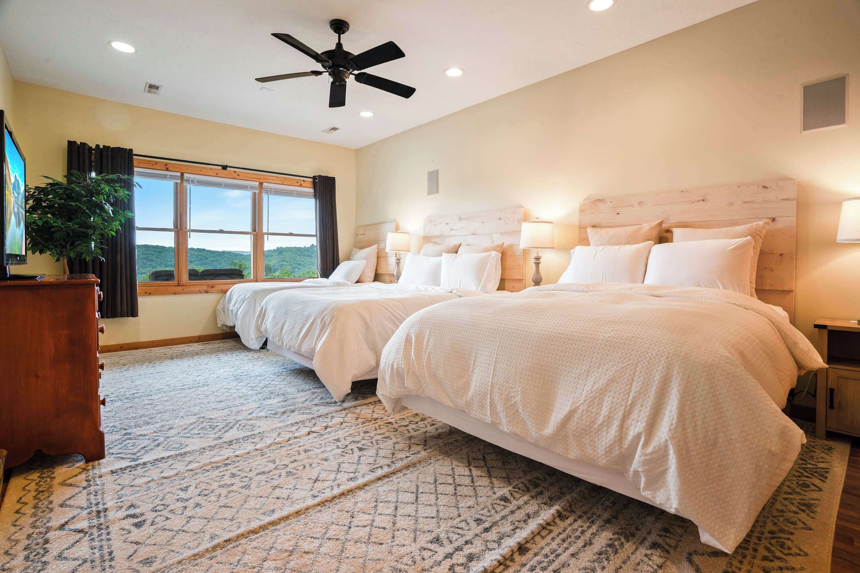 A bedroom with two beds, a ceiling fan, a large window with a view, and a patterned rug.
