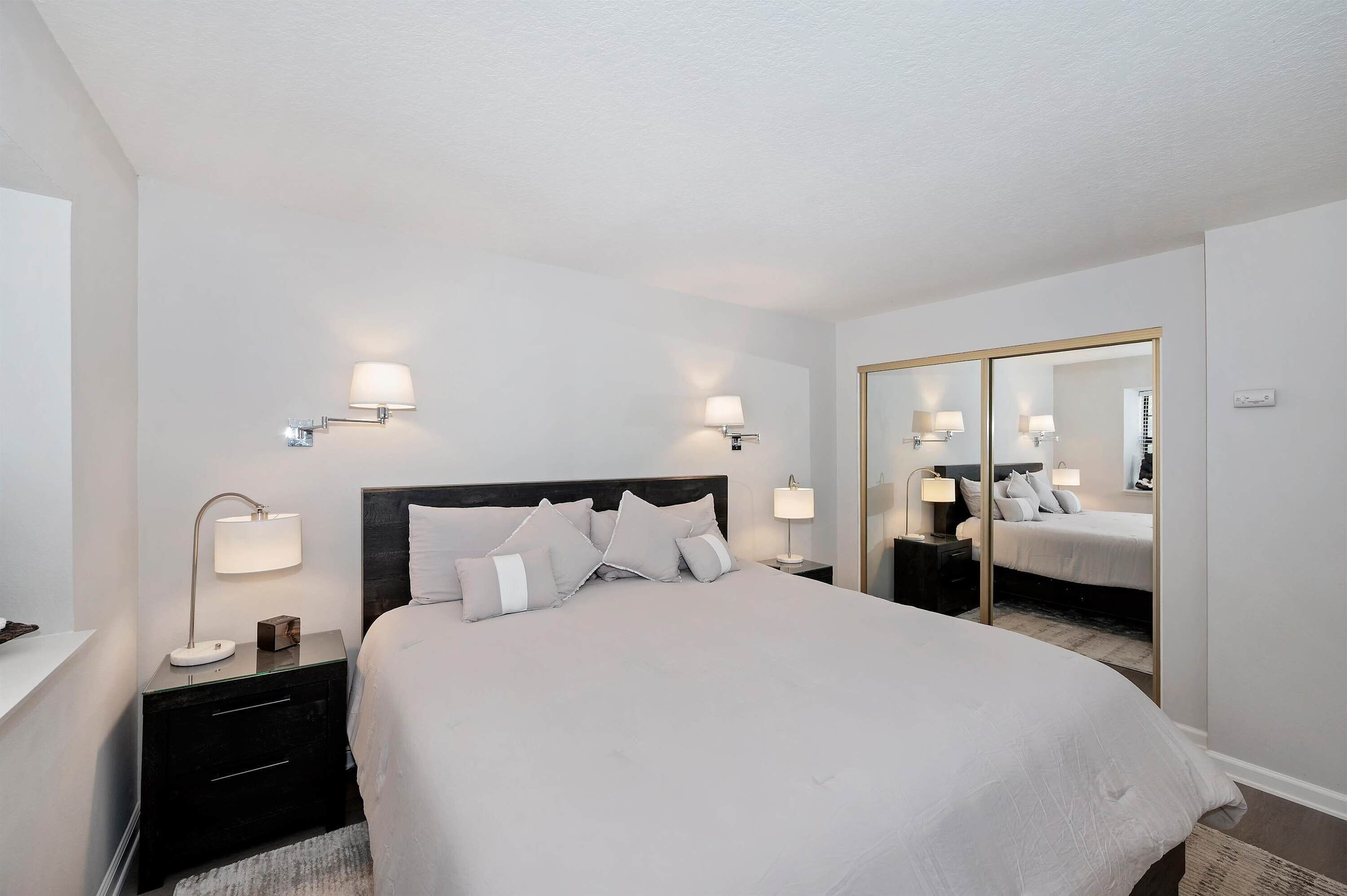 A hotel room with a bed, bedside lamps, a nightstand, a mirror, and a wardrobe.