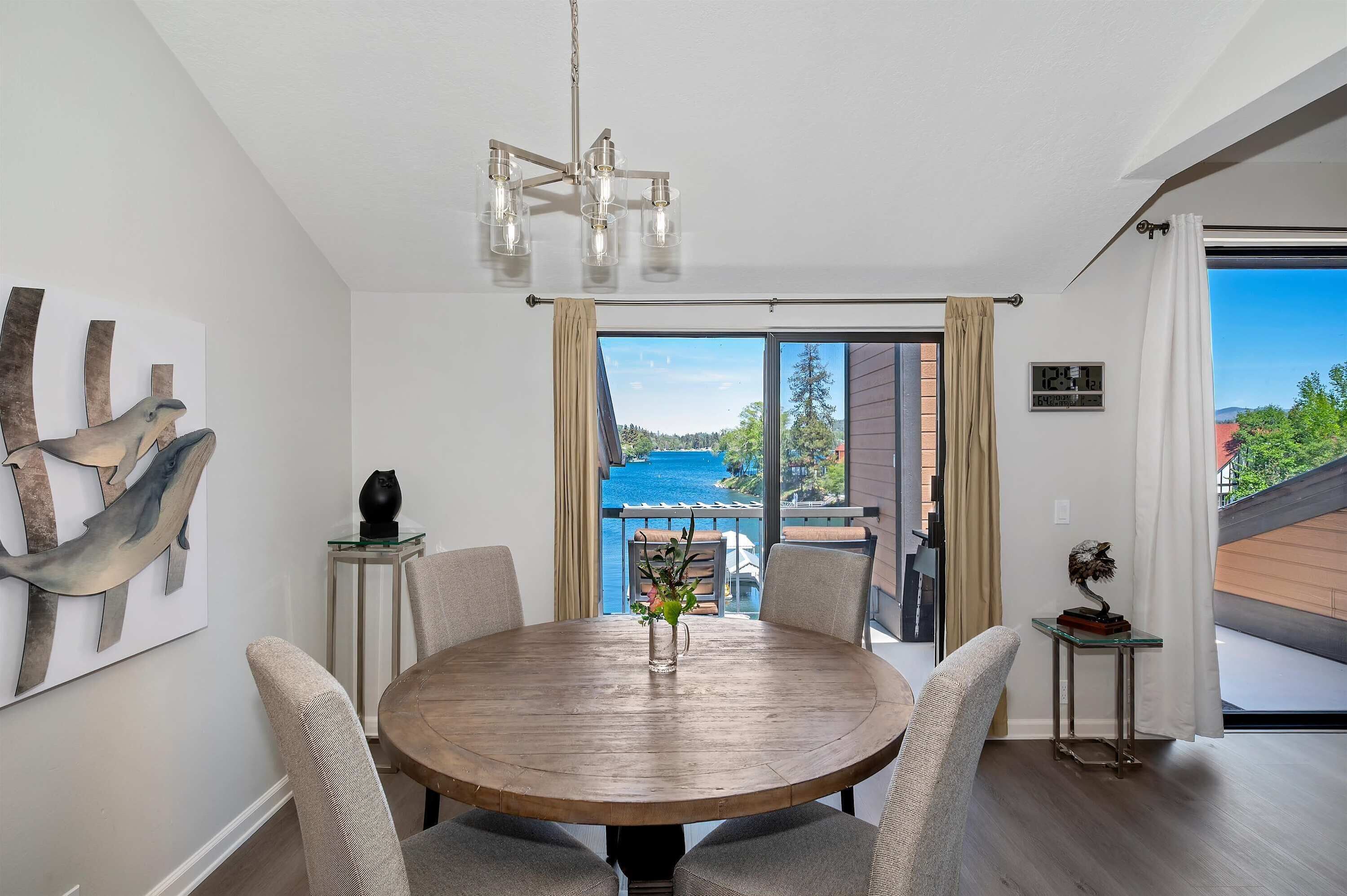 A dining area with a round wooden table, chairs, a chandelier, and a view of a blue sky and greenery through a large window.