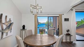 A dining area with a round wooden table, chairs, a chandelier, and a view of a blue sky and greenery through a large window.