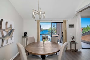 A dining area with a round wooden table, chairs, a chandelier, and a view of a blue sky and greenery through a large window.