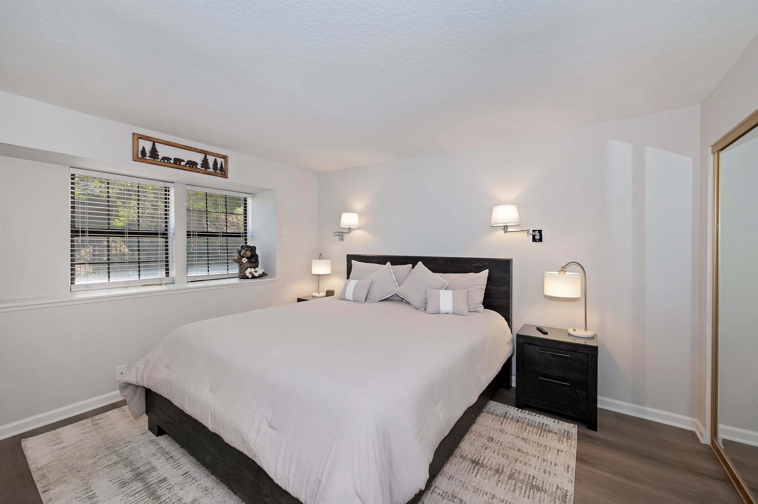 A bedroom with a large bed, two bedside tables with lamps, a window with blinds, and a wall-mounted picture.