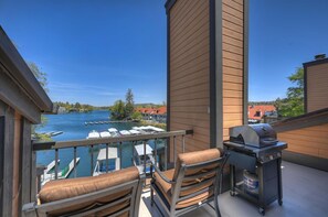 A balcony with a grill, chairs, and a view of a lake with boats.