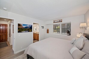 A bedroom with a large bed, a window with blinds, a wall-mounted lamp, and a door leading to another room.