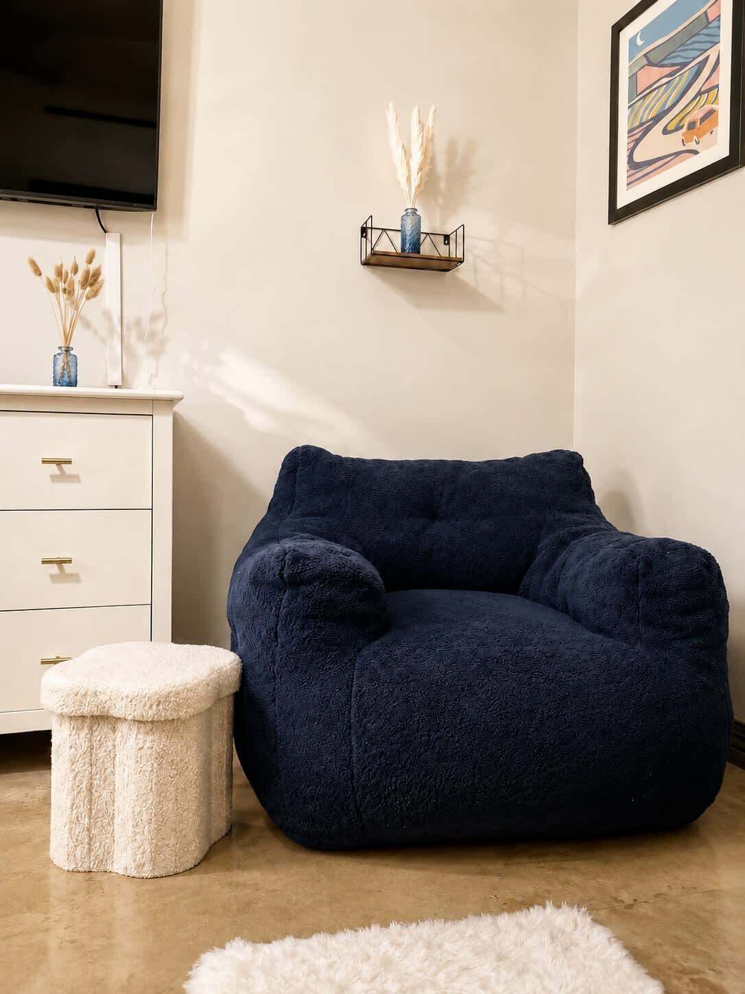 Ein gemütliches Zimmer mit einem dunkelblauen Sitzsack, einer weißen Kommode mit goldfarbenen Griffen, einem kleinen weißen Hocker und einem gerahmten abstrakten Gemälde an der Wand.