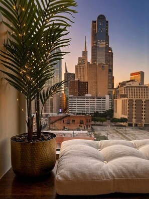 A cityscape view from a window with a potted plant and a cushioned seating area. - Skyline Views | Valet Parking | Resort Amenities (Dallas)