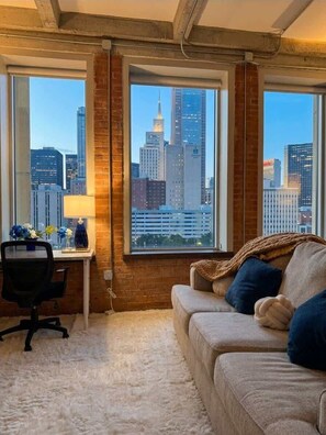 A room with a city view, a desk with a lamp, a chair, and a sofa with a blanket. - Skyline Views | Valet Parking | Resort Amenities (Dallas)
