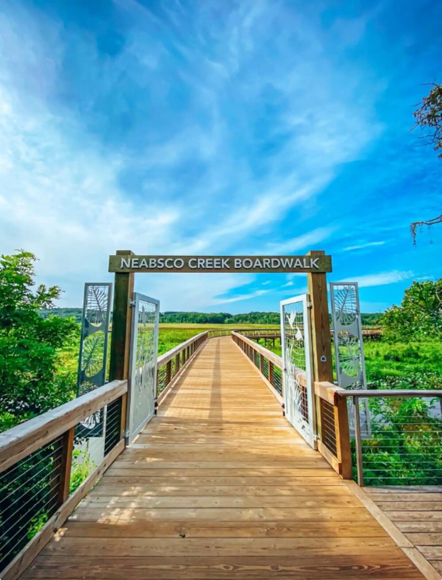 Une promenade en bois, agrémentée d’un panneau indiquant « NEABSC CO CREEK BOARDWALK », qui mène à un paysage naturel.