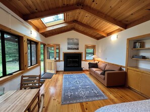 A cozy living room with wooden floors, a fireplace, a sofa, a dining table with chairs, and large windows.