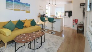 A modern living room with a yellow sofa, green and yellow cushions, a coffee table, and a kitchen area with a dining table and bar stools.
