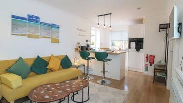 Uma sala de estar moderna com um sofá amarelo, almofadas verdes e amarelas, uma mesa de centro e uma área de cozinha com uma mesa de jantar e banquetas.