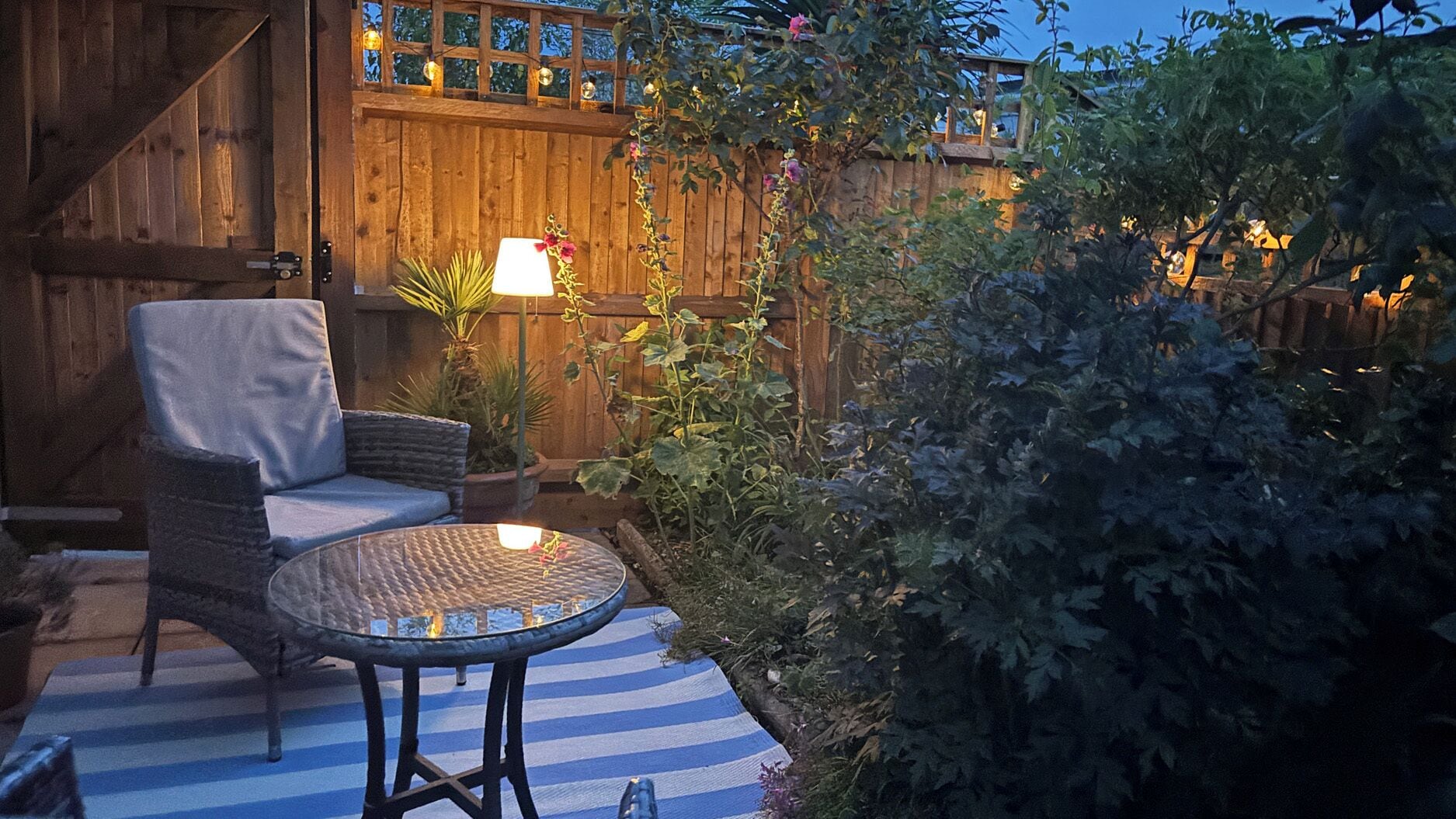 A patio with a round table and a cushioned chair, surrounded by a wooden fence and lush greenery.