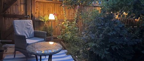 A patio with a round table and a cushioned chair, surrounded by a wooden fence and lush greenery.