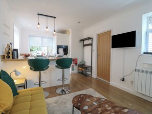 Un espacio habitable compacto con kitchenette, una zona de comedor con dos taburetes de bar, un sofá y un televisor montado en la pared.