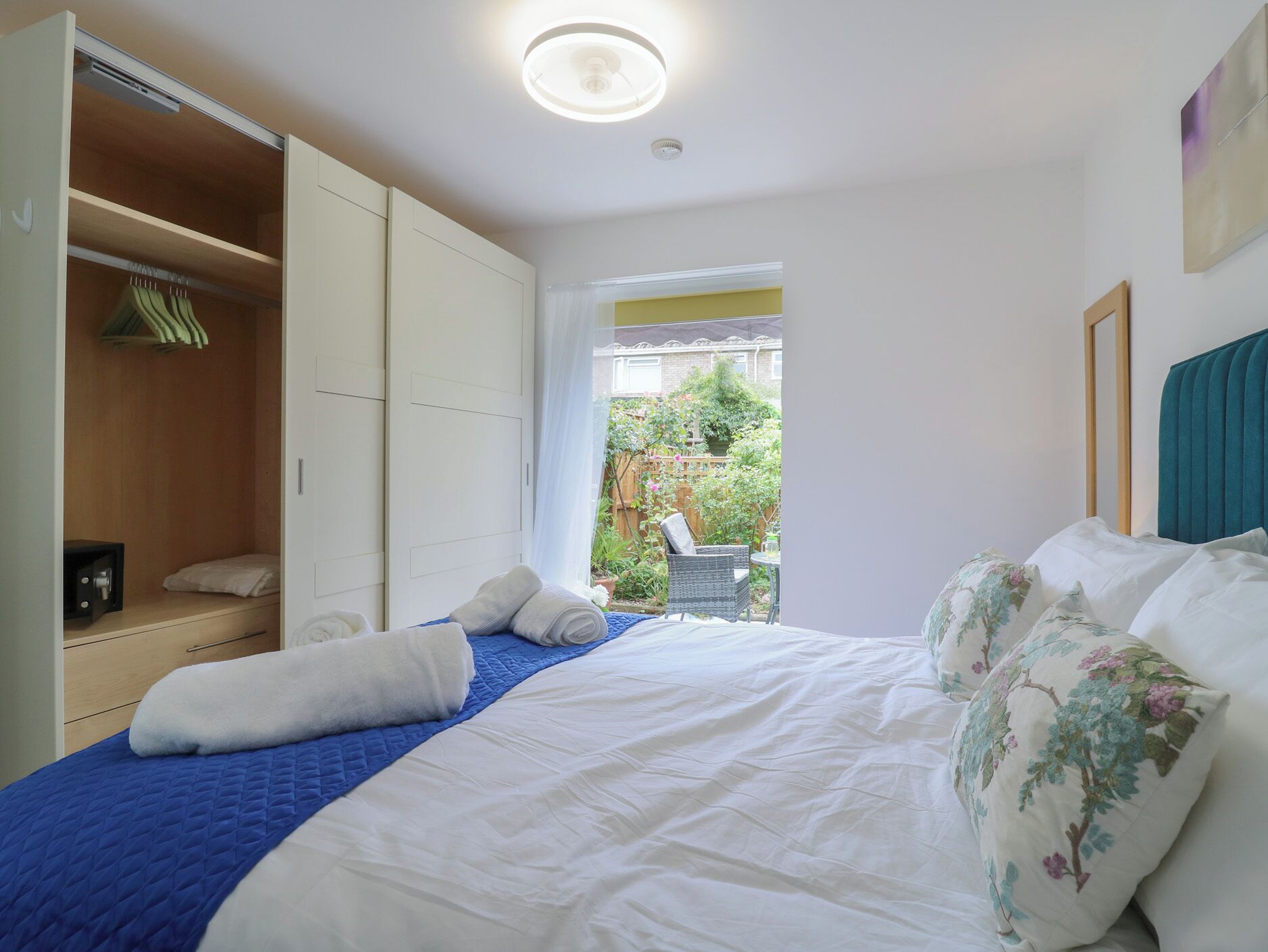 A bedroom with a bed, a wardrobe, a small table, and a view of a garden.