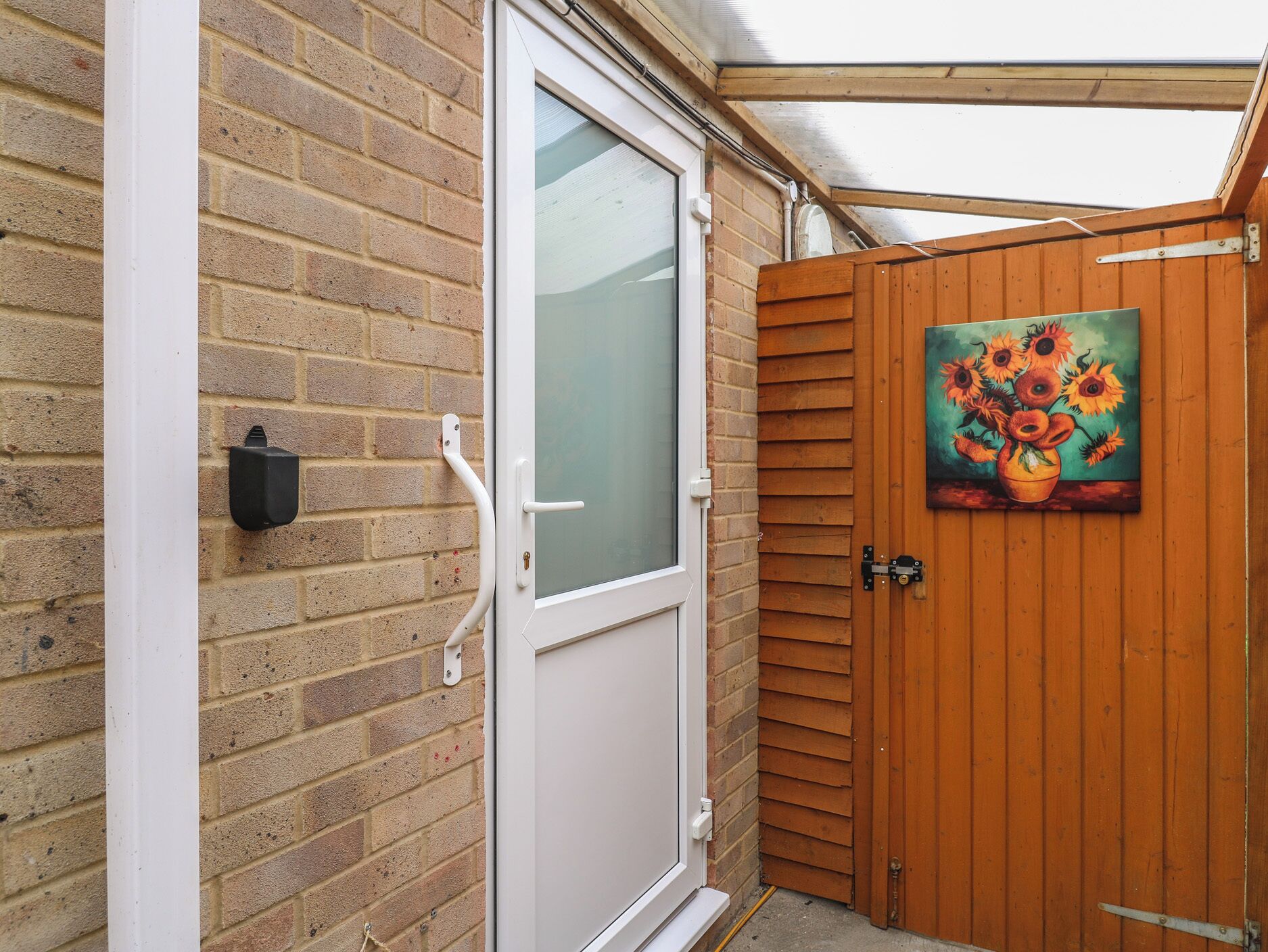 A white door with a glass panel, a brick wall, a wooden gate, and a painting of sunflowers.
