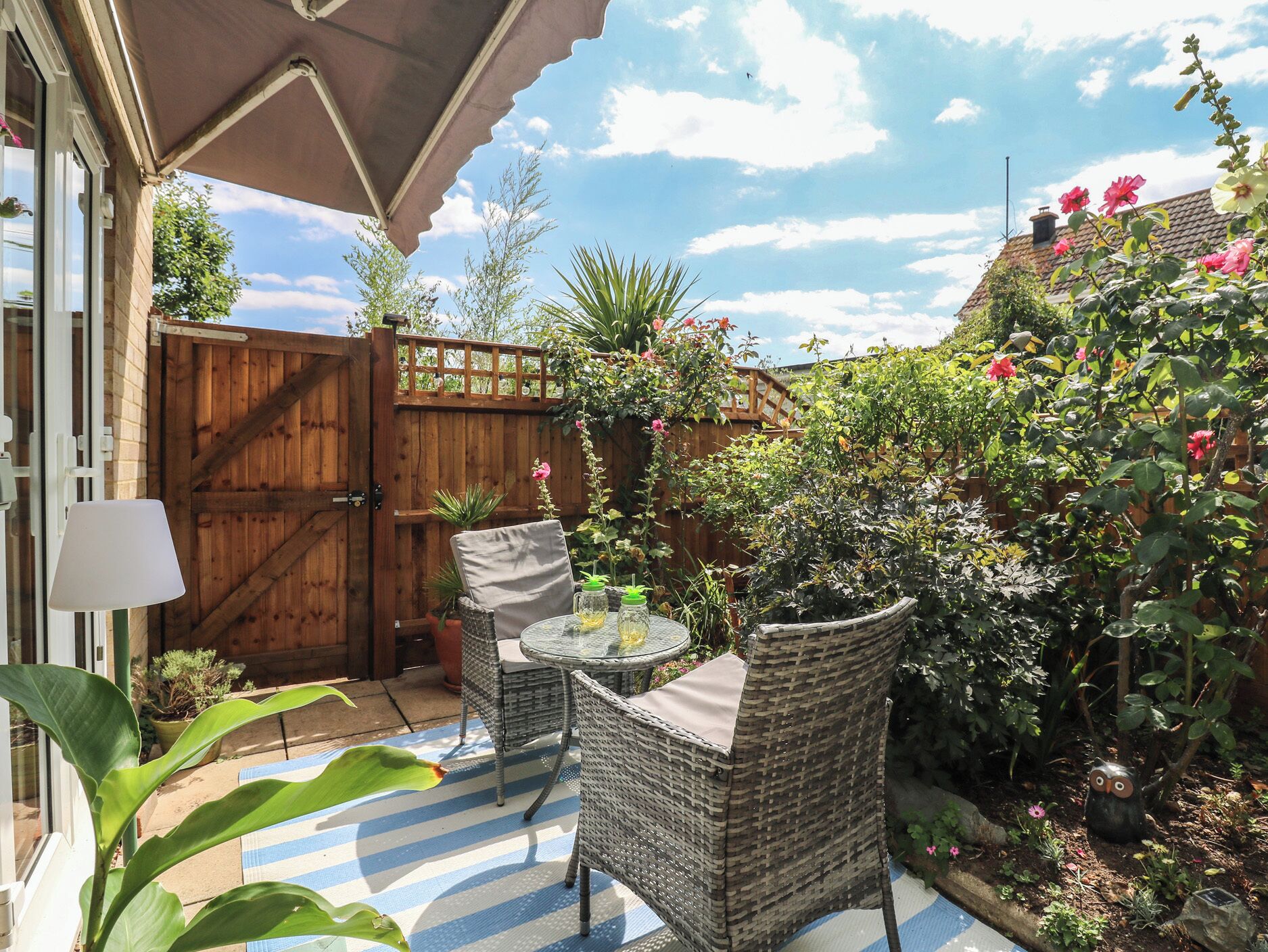 A patio with wicker furniture, a table, and chairs, surrounded by a wooden fence and various plants.