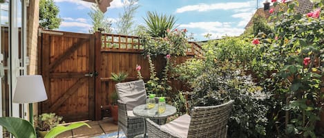 Une terrasse dotée de meubles en osier, d’une table et de chaises, entourée d’une clôture en bois et agrémentée de diverses plantes.