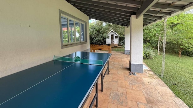 Ett bordtennisbord under ett takförsett område med ett stengolv, ett fönster och en liten träkonstruktion i bakgrunden.