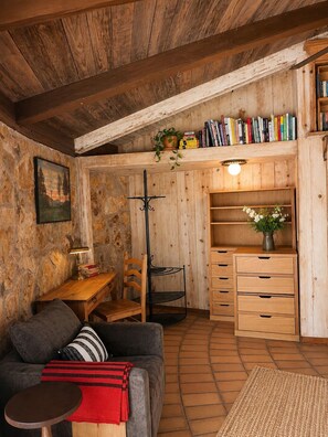 Ein gemütliches Zimmer mit Holzbalkendecke, einem Bücherregal, einem Schreibtisch, einem Sessel, einem Sofa und einem kleinen Tisch.