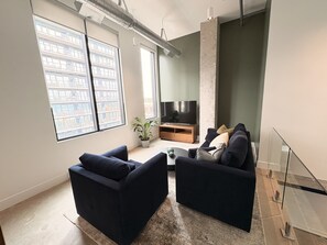 A modern living room with a large window, a flat-screen TV, dark sofas, and a coffee table. - 1 BR Industrial-Chic Penthouse Loft | City Views (Halifax)