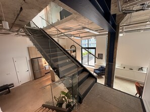 Um interior moderno com uma escada de vidro que desce para um nível inferior, uma área de estar com vista para o exterior e dutos expostos no teto.