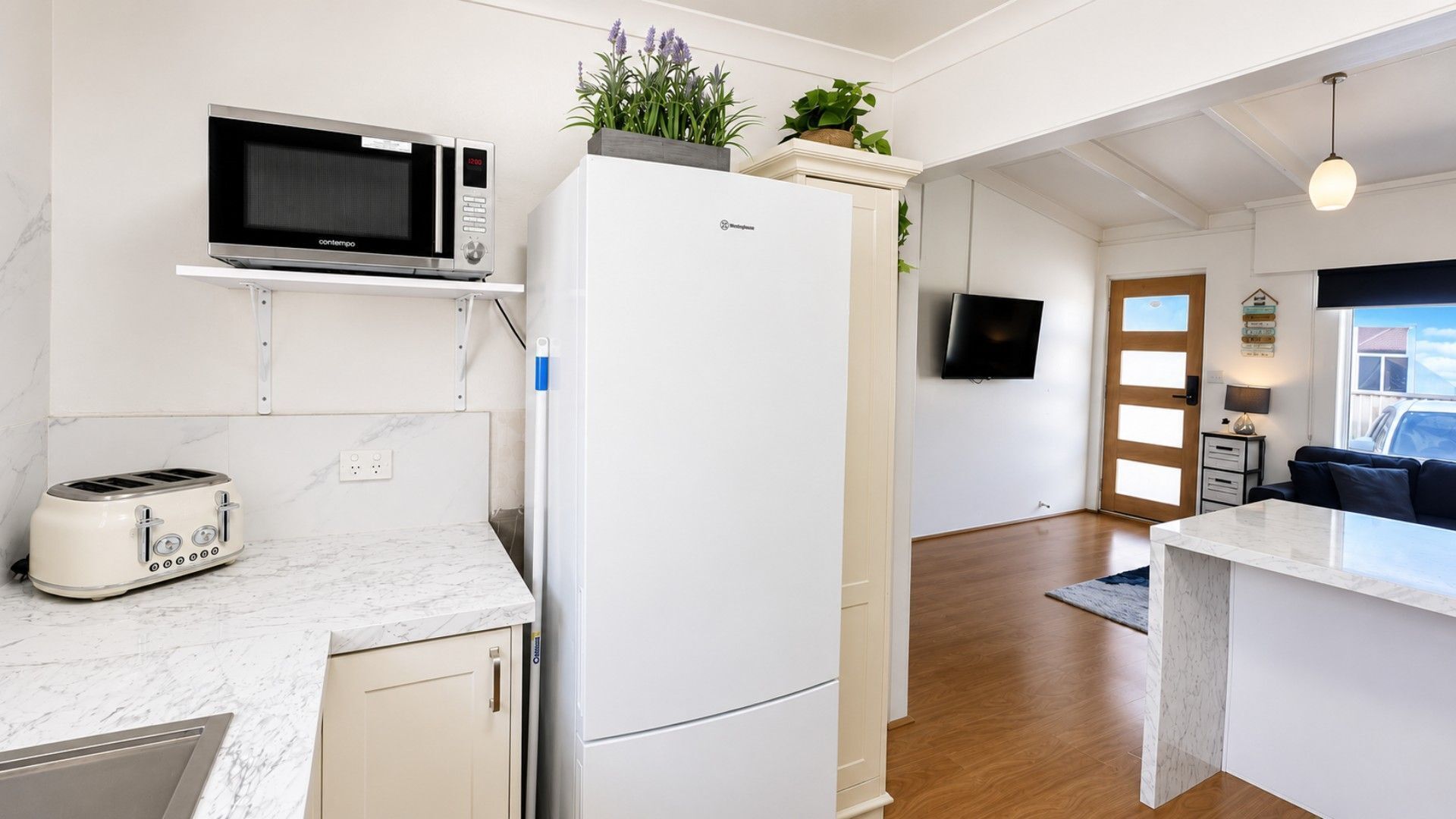 A compact kitchen with a microwave, toaster, and refrigerator, a television mounted on the wall, and a view of the outdoors through a window.