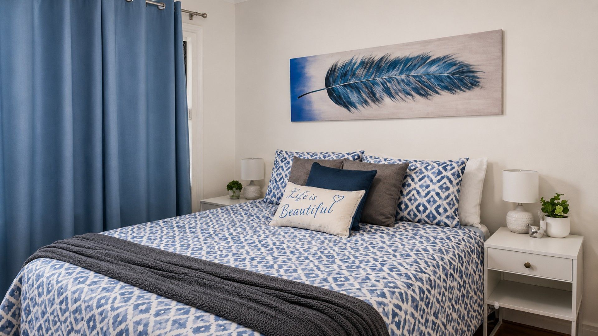 A bedroom with a patterned bedspread, pillows, a blue curtain, a wall-mounted painting, bedside tables with lamps, and a small plant.