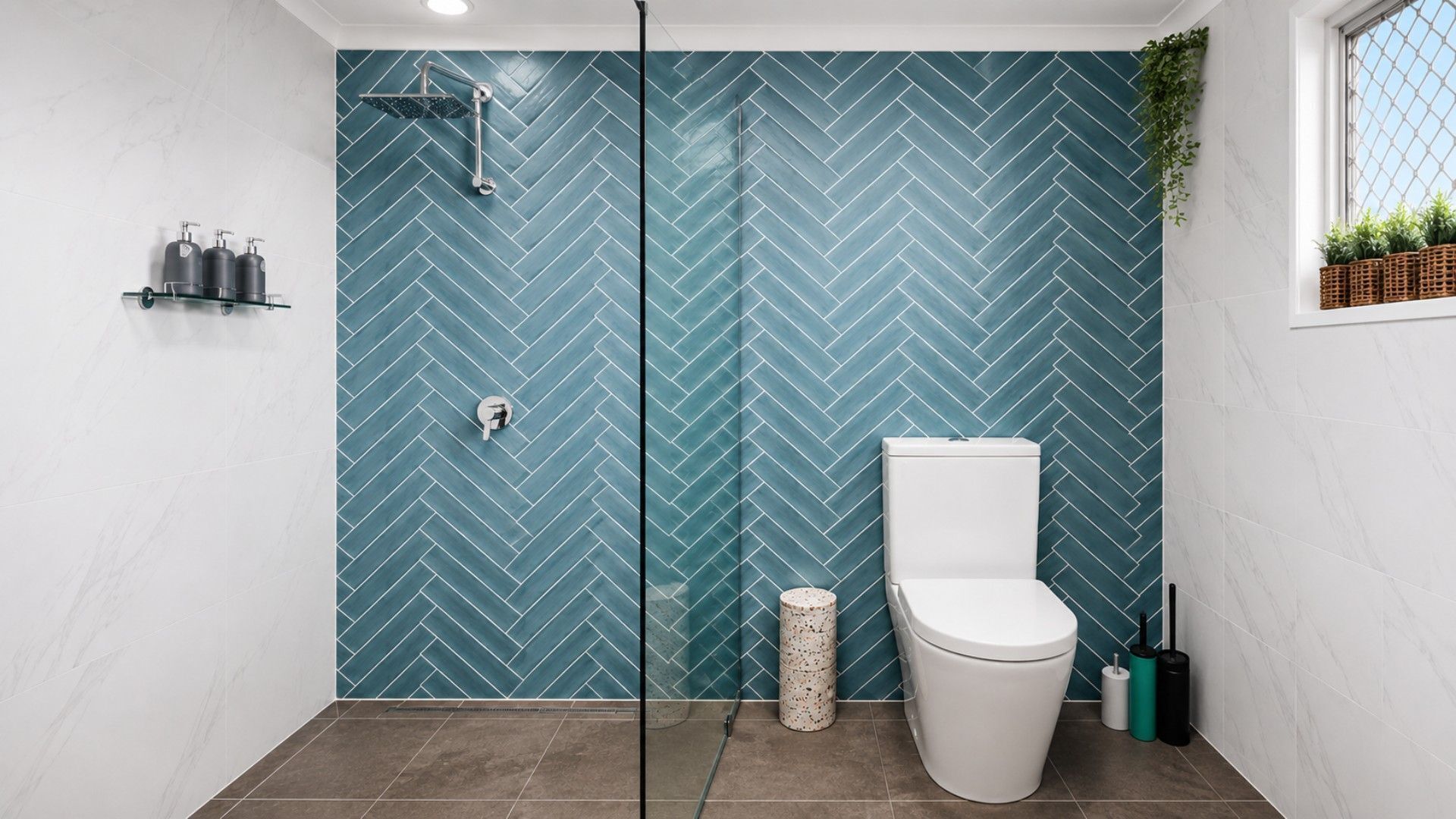 Une salle de bain avec un carrelage disposé en motif «chevrons», une douche en verre, des toilettes et une petite plante posée sur le rebord de la fenêtre.