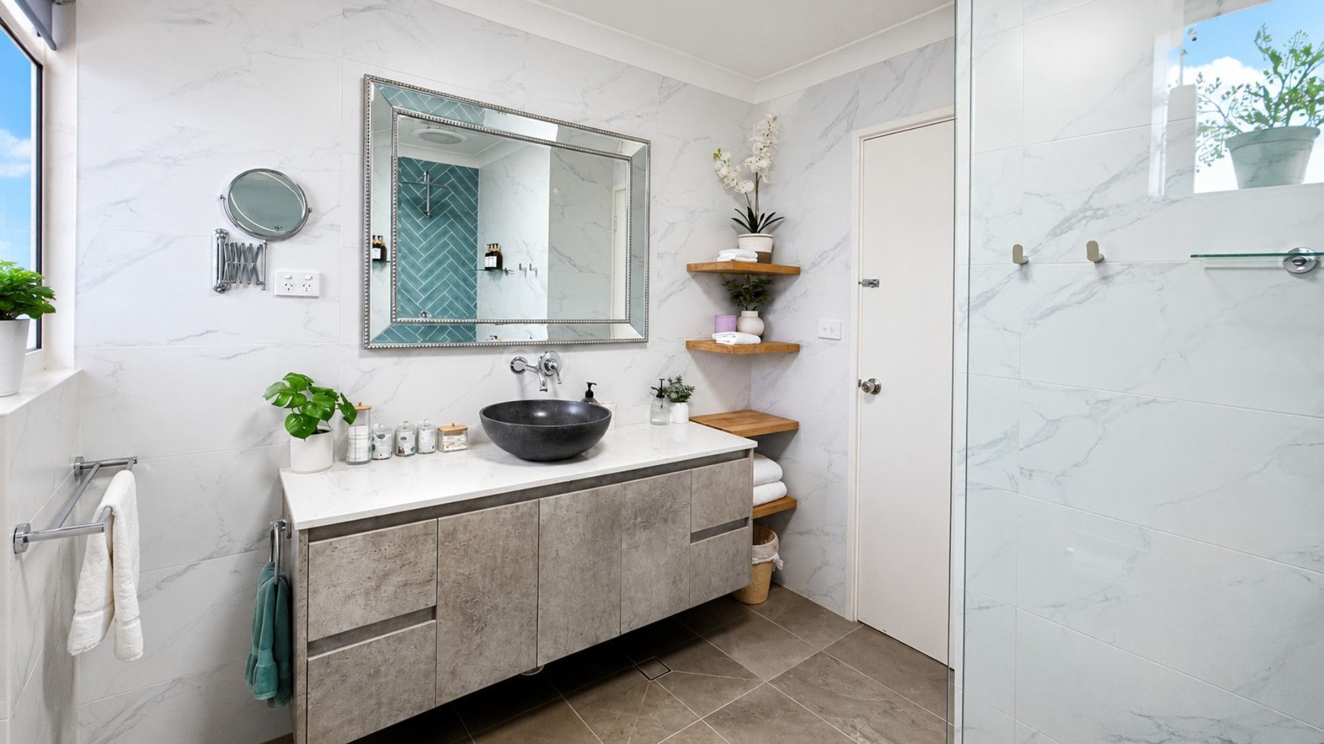 A modern bathroom with a large mirror, a black vessel sink, marble tiles, and a glass shower enclosure.